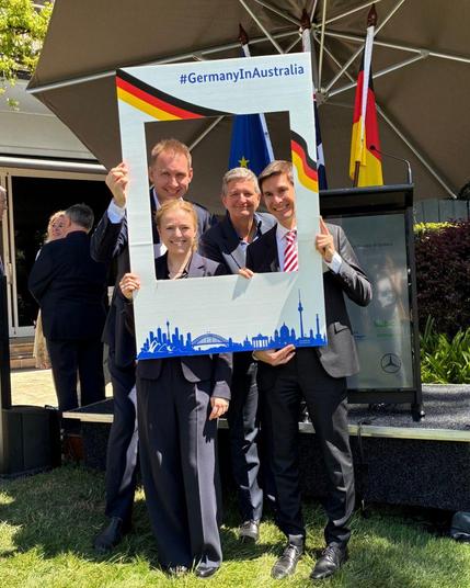 Gruppenfoto vom Team BSI bei den Feierlichkeiten zum Tag der Deutschen Einheit in Australien. V.l.n.r. Lara Riefle, Michael Dwucet, BSI-Präsidentin Claudia Plattner, Florian Thurner. Sie halten einen Polaroid-Rahmen in den Händen.